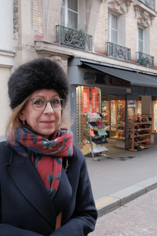 Dominique · 10/01/2026 · 1333 x 2000 Cette photographie met en scène une femme comme sujet principal, vêtue d’un manteau sombre, d’un foulard coloré et d’un chapeau en fourrure, avec des lunettes. Le cadrage est serré sur elle, avec un arrière-plan flou montrant une boutique et une rue parisienne.La lumière est naturelle et douce, typique d’un jour couvert. Les couleurs dominantes sont les tons neutres (gris, noir) adoucis par les touches vives du foulard (rouge, orange) et les détails architecturaux (bois, fer forgé).L’ambiance est urbaine et élégante, évoquant une atmosphère calme et intemporelle.