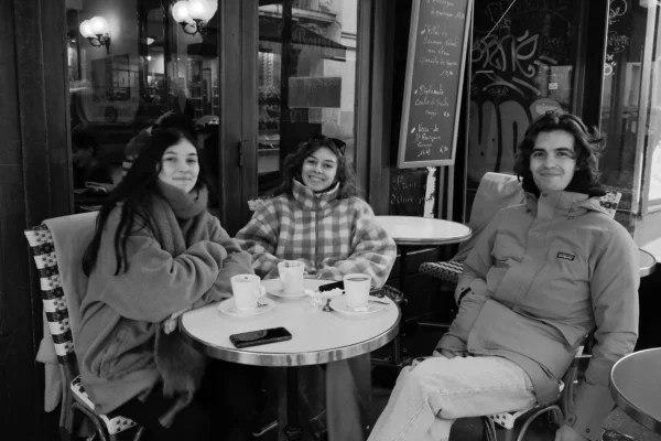 Dominique · 10/01/2026 · 2000 x 1333 Cette photographie en noir et blanc montre trois personnes assises autour d'une table dans un café. Le sujet principal est leur interaction conviviale.La composition est symétrique, avec les trois individus placés en triangle autour de la table. Le cadrage est serré, centré sur eux et leur environnement immédiat.La lumière est douce et naturelle, probablement artificielle, éclairant le sujet sans créer d'ombres marquées. Les couleurs dominantes, bien que en noir et blanc, suggèrent des tons clairs et contrastés.Le style est documentaire et intime, capturant un moment de vie quotidienne. L'ambiance est chaleureuse et détendue.