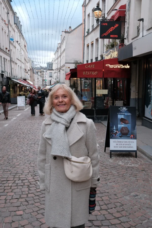 Dominique · 10/01/2026 · 1333 x 2000 Cette photographie met en scène une femme âgée comme sujet principal, souriante, vêtue d’un long manteau beige et d’un chapeau assorti. La composition montre une rue pavée animée, avec des boutiques et des passants en arrière-plan.Le cadrage est serré sur la femme, mais inclut suffisamment d’éléments urbains pour situer le contexte. La lumière est naturelle, douce, probablement en journée, avec des ombres discrètes. Les couleurs dominantes sont les tons neutres (beige, gris) et quelques touches de rouge et de vert des enseignes.L’ambiance est chaleureuse et paisible, évoquant une balade urbaine tranquille.