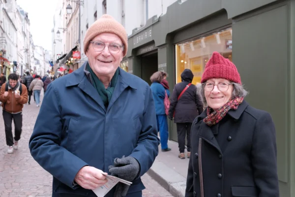 Dominique · 10/01/2026 · 2000 x 1333 Cette photographie montre deux personnes âgées comme sujets principaux, souriantes et posant côte à côte. La composition est simple et naturelle, avec un cadrage serré sur eux, tout en laissant voir un fond légèrement flou de passants et de bâtiments.La lumière semble naturelle, probablement en extérieur par temps couvert. Les couleurs dominantes sont sobres : tons gris et bleus pour les vêtements, avec des touches chaudes comme le rouge du bonnet de la femme. Le style est documentaire et spontané, créant une ambiance chaleureuse et conviviale.