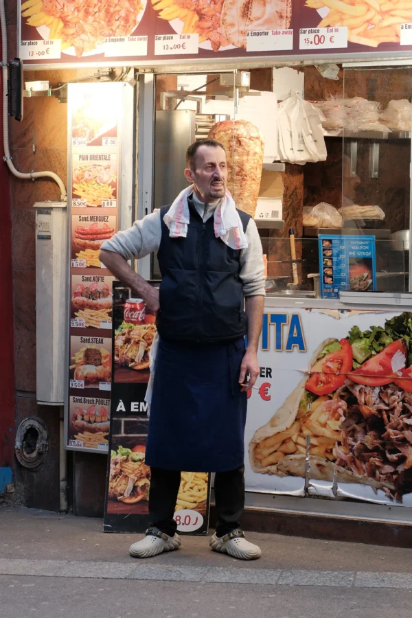 Dominique · 10/01/2026 · 1333 x 2000 Cette photographie montre un homme en tablier de cuisine, sujet principal, posant devant un stand de pizzas. La composition met en valeur son attitude décontractée, un bras sur la hanche. Le cadrage est serré, centré sur lui et son environnement immédiat.La lumière semble naturelle, légèrement tamisée, créant des ombres douces. Les couleurs dominantes sont chaudes : tons beiges, rouges et blancs des enseignes et des aliments. L’ambiance est conviviale et artisanale, évoquant un marché ou une rue animée. Le style est réaliste et spontané.