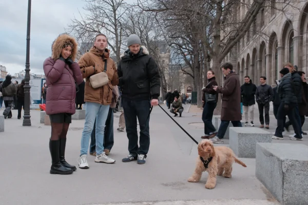 Dominique · 10/01/2026 · 2000 x 1333 Cette photographie capture un groupe de personnes en ville, dont le sujet principal est un chien assis sur le trottoir, tenu en laisse. La composition montre une scène urbaine avec des passants en arrière-plan.Le cadrage est serré sur le chien et ses propriétaires, tout en incluant l’environnement. La lumière est naturelle, probablement hivernale, avec des ombres marquées. Les couleurs dominantes sont neutres et froides : gris, beige et marron, adoucies par les vêtements chauds des personnes.L’ambiance est calme et quotidienne, reflétant une scène de vie urbaine en hiver.