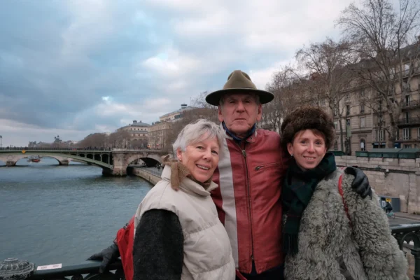Dominique · 10/01/2026 · 2000 x 1333 Cette photographie montre trois personnes posant ensemble devant un pont et une rivière. Le sujet principal est ce trio souriant, composé d’un homme au centre avec un chapeau et deux femmes de chaque côté.La composition est symétrique et harmonieuse, avec le pont en arrière-plan. Le cadrage est serré sur les sujets, mais inclut aussi l’environnement urbain. La lumière est naturelle, légèrement voilée par un ciel nuageux, créant une ambiance douce et sobre. Les couleurs dominantes sont les tons neutres (gris, beige) et quelques touches de rouge et blanc dans les vêtements. Le style est documentaire et chaleureux, évoquant une ambiance détendue et conviviale.