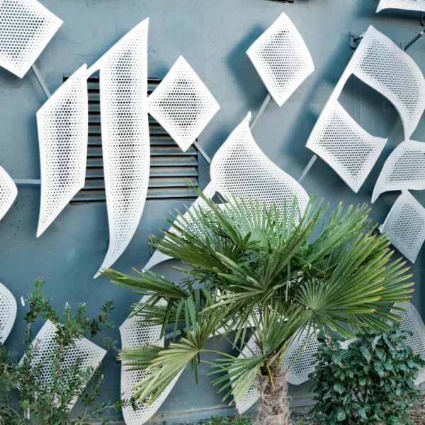 Nathalie F. · 17/01/2026 · 4160 x 4160 Cette photographie met en scène une installation artistique murale composée de pages de livres disposées en motifs géométriques sur un fond bleu. Le sujet principal est cette structure textuelle, contrastant avec des plantes vertes en bas, dont un palmier et des fougères.La composition est équilibrée, avec un cadrage serré sur le mur et les végétaux. La lumière naturelle éclaire uniformément la scène, mettant en valeur les textures et les couleurs. Les teintes dominantes sont le bleu grisâtre du mur, le blanc des pages et le vert des plantes. Le style est épuré et contemporain, créant une ambiance à la fois poétique et intellectuelle, mêlant nature et culture.