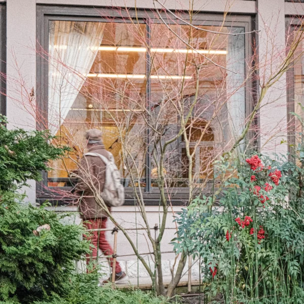 Nathalie F. · 17/01/2026 · 4160 x 4160 Cette photographie montre une personne en mouvement, vêtue d’un manteau clair et d’un pantalon rouge, traversant un jardin devant une fenêtre. Le sujet principal est flou, suggérant un mouvement rapide.La composition utilise un cadrage serré avec des éléments naturels en premier plan : branches nues et plantes. La lumière est douce, naturelle, avec des ombres discrètes. Les couleurs dominantes sont les tons froids (gris, vert pâle) et chauds (rouge, orange des feuilles et du manteau).Le style est réaliste et spontané, évoquant une ambiance calme et légèrement mélancolique, renforcée par les branches sans feuilles.