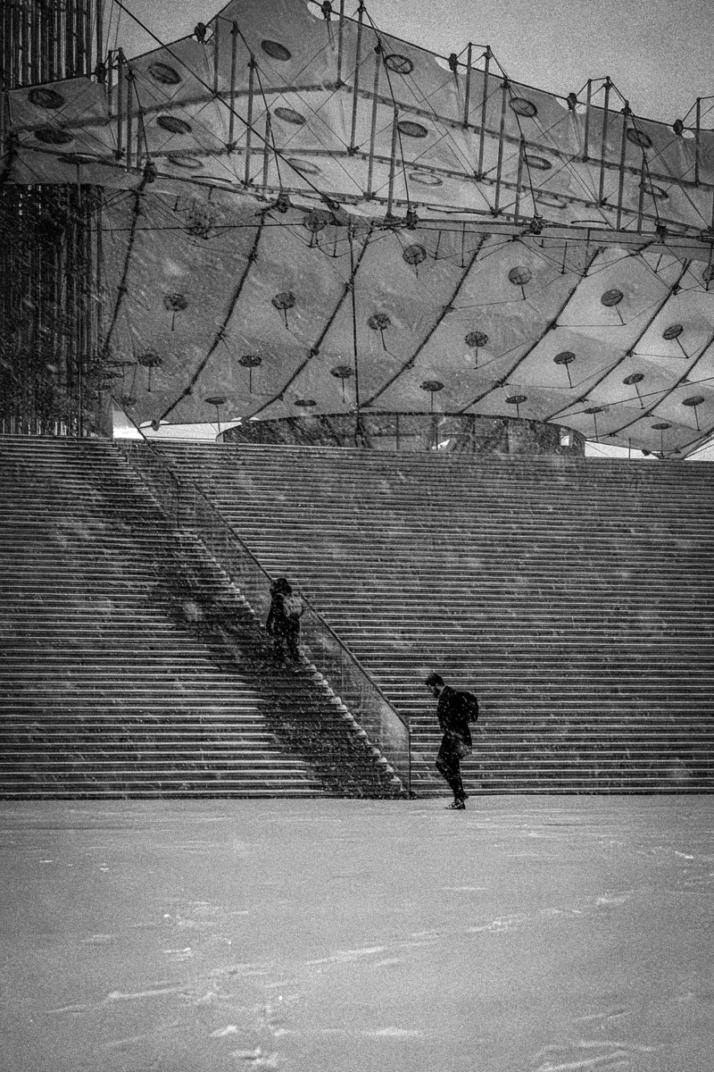AnJo · 07/01/2026 · 1280 x 1920 Cette photographie en noir et blanc capture deux enfants skiant sur une pente enneigée. Le sujet principal est leur mouvement dynamique, mis en valeur par le cadrage serré et les lignes des marches en arrière-plan.La composition utilise les gradins comme élément structurant, créant une perspective profonde. La lumière, douce et diffuse, adoucit les contrastes, renforçant l’ambiance hivernale et nostalgique. Les tons gris et blancs dominent, accentuant le côté épuré et intemporel.Le style, sobre et documentaire, évoque une scène authentique et spontanée, imprégnée de simplicité et de joie enfantine.