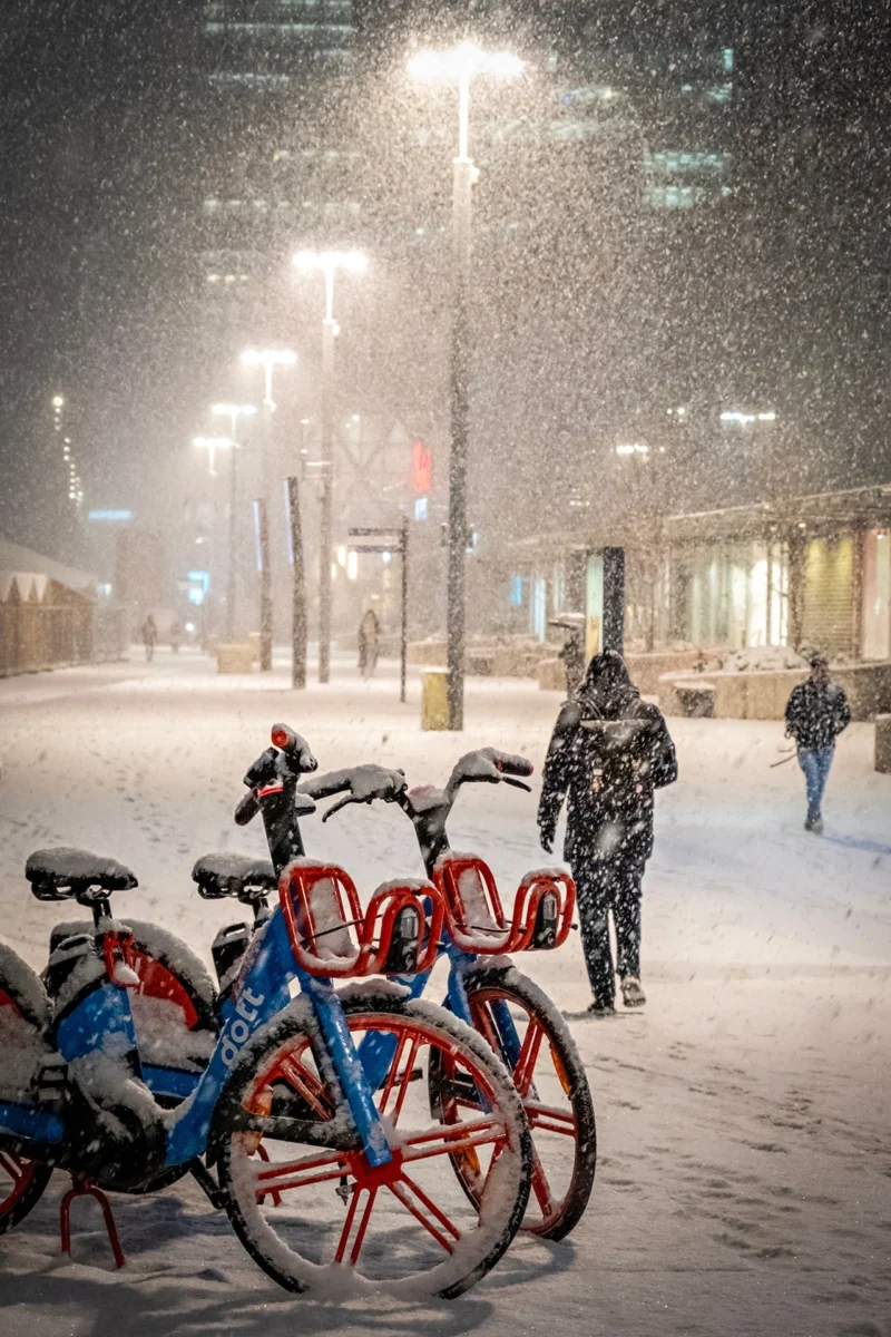 AnJo · 07/01/2026 · 1280 x 1920 Cette photographie capture une scène hivernale urbaine. Le **sujet principal** est un groupe de vélos en libre-service, partiellement recouverts de neige, avec deux personnes en arrière-plan.La **composition** joue sur la perspective, avec un cadrage serré sur les vélos et une profondeur de champ qui inclut les bâtiments et les passants. La **lumière**, douce et tamisée, provient des réverbères, créant une ambiance nocturne et feutrée.Les **couleurs dominantes** sont les tons froids : blanc de la neige, bleu des vélos, et gris des bâtiments. Le style est **réaliste et documentaire**, évoquant une **ambiance calme et mélancolique**, typique d’une nuit d’hiver en ville.