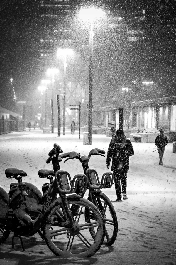 AnJo · 07/01/2026 · 1280 x 1920 Cette photographie en noir et blanc capture une scène nocturne enneigée. Le sujet principal est un vélo abandonné, au premier plan, avec des personnes en arrière-plan marchant sur un trottoir. La composition joue sur les contrastes entre l'avant et l'arrière-plan, créant une profondeur.Le cadrage est serré sur le vélo, mais inclut aussi des éléments urbains comme les lampadaires et les bâtiments. La lumière est douce, diffusée par les réverbères, créant une ambiance tamisée et poétique. Les couleurs dominantes sont les nuances de gris, renforçant l'atmosphère hivernale et mélancolique. Le style est documentaire et évocateur, évoquant une touche de nostalgie.