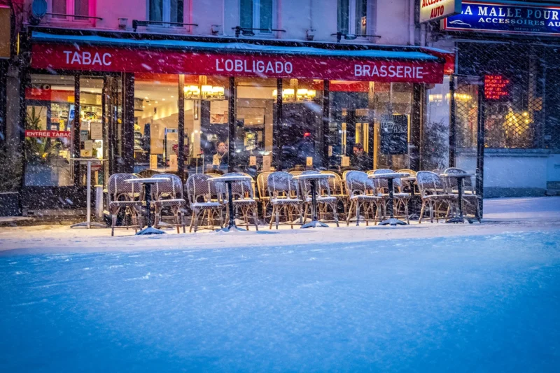 AnJo · 07/01/2026 · 1920 x 1280 Cette photographie montre une terrasse de café parisienne enneigée, nommée **« L'Obligatoire »**, avec des tables et chaises alignées sous une bâche. Le sujet principal est cette terrasse déserte, contrastant avec l’animation intérieure éclairée.La composition est symétrique, avec un cadrage serré sur la terrasse et un premier plan de neige. La lumière, douce et artificielle, provient des enseignes et vitrines, créant une ambiance chaleureuse malgré le froid. Les couleurs dominantes sont les tons froids (blanc de la neige, bleu nuit) et chauds (rouge des enseignes, lumières jaunes).Le style est urbain et nocturne, évoquant une ambiance poétique et mélancolique.