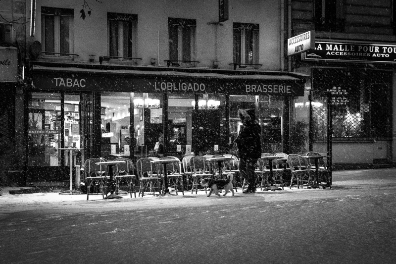 AnJo · 07/01/2026 · 1920 x 1280 Cette photographie en noir et blanc capture une terrasse de café nocturne. Le sujet principal est l’ambiance animée des clients attablés, éclairés par des lumières douces et tamisées. La composition met en valeur les tables et chaises disposées devant un magasin de tabac et un bar à vin, avec des enseignes lumineuses.Le cadrage est serré, centré sur la terrasse et ses occupants. La lumière, artificielle et chaleureuse, crée des contrastes doux. Les couleurs dominantes, bien que monochromes, évoquent des tons gris et noirs avec des touches de blanc lumineux.Le style est documentaire et intemporel, évoquant une ambiance paisible et conviviale, typique d’un quartier urbain la nuit.
