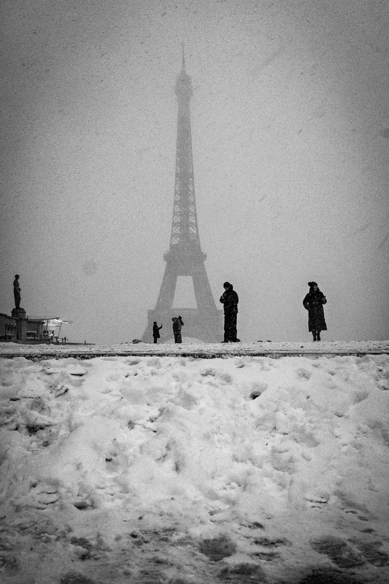 AnJo · 07/01/2026 · 1280 x 1920 Cette photographie en noir et blanc met en scène la **Tour Eiffel**, sujet principal, partiellement voilée par le brouillard. La composition montre des silhouettes de personnes marchant sur un pont ou une berge enneigée, ajoutant une touche d’échelle et de vie.Le cadrage est serré, centré sur la tour et les silhouettes, avec une perspective légèrement basse. La lumière est diffuse, atténuée par la brume, créant une ambiance mystérieuse et mélancolique. Les tons dominants sont les gris et noirs, renforçant le côté dramatique et poétique. Le style est documentaire et artistique, évoquant une atmosphère froide et contemplative.