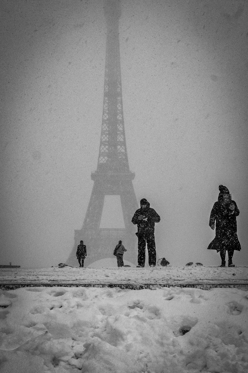 AnJo · 07/01/2026 · 1280 x 1920 Cette photographie en noir et blanc met en scène la **Tour Eiffel**, sujet principal, partiellement dissimulée par le brouillard. La **composition** place la tour au centre, avec des silhouettes de personnes en premier plan, ajoutant une échelle humaine.Le **cadrage** est serré, centré sur les visiteurs et la base de la tour. La **lumière** est diffuse, atténuée par la brume, créant une atmosphère mystérieuse. Les **couleurs dominantes** sont les gris et noirs, renforçant le côté dramatique.Le **style** est documentaire et poétique, évoquant une **ambiance** froide et mélancolique, typique d’un hiver parisien.