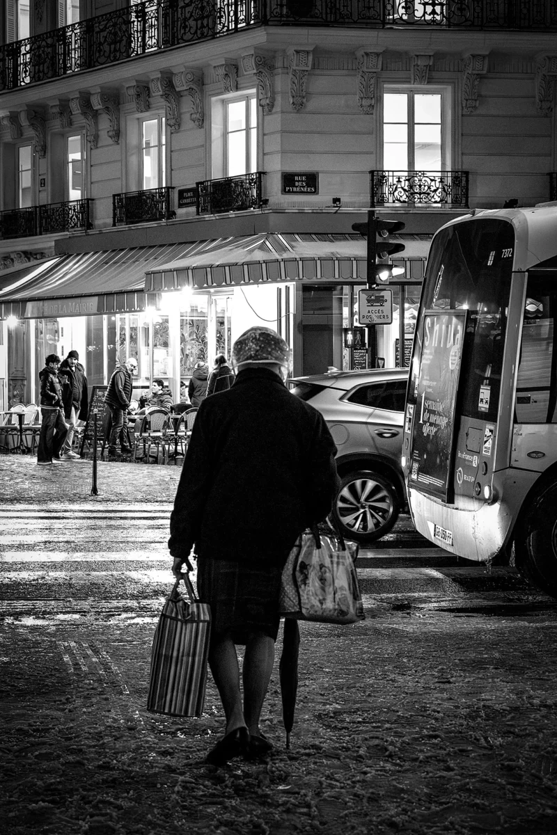 AnJo · 07/01/2026 · 1280 x 1920 Cette photographie en noir et blanc met en scène une personne âgée, vue de dos, marchant avec des sacs de courses. Le sujet principal est centré, avec une composition équilibrée.Le cadrage serré isole le personnage, tout en intégrant l’arrière-plan urbain. La lumière douce, tamisée, crée une ambiance nocturne et mélancolique. Les contrastes entre ombres et clartés accentuent les détails architecturaux et les silhouettes.Les tons dominants, du noir au gris clair, renforcent l’atmosphère intemporelle et poétique de la scène, évoquant une ville paisible et calme.