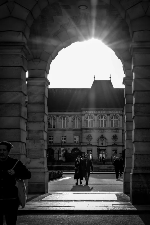 AnJo · 17/01/2026 · 1280 x 1920 Cette photographie en noir et blanc capture un passage architectural avec une arche centrale. Le sujet principal est le soleil bas, créant un fort contraste lumineux et des rayons traversant l’arche. La composition est symétrique, avec des colonnes de chaque côté encadrant la scène.Le cadrage est serré, mettant en valeur les silhouettes des personnes et les bâtiments historiques en arrière-plan. La lumière dramatique, avec ses contrastes marqués, accentue l’ambiance mystérieuse et solennelle. Les tons dominants sont les noirs profonds et les blancs éclatants, typiques du style photographique noir et blanc classique, évoquant une atmosphère à la fois élégante et mélancolique.