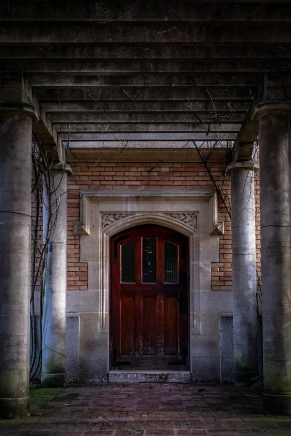 AnJo · 17/01/2026 · 1280 x 1920 Cette photographie met en scène une porte en bois rouge sombre, sujet principal, encadrée par une architecture en brique et pierre vieillissante. La composition est symétrique, avec un cadrage serré qui accentue le passage étroit et l’architecture gothique.La lumière, tamisée et contrastée, crée une ambiance mystérieuse et dramatique. Les couleurs dominantes sont les tons grisâtres des murs, les briques rouges et le bois foncé de la porte. Le style évoque un réalisme sombre, presque cinématographique. L’ambiance est mélancolique et envoûtante, renforcée par l’obscurité environnante.