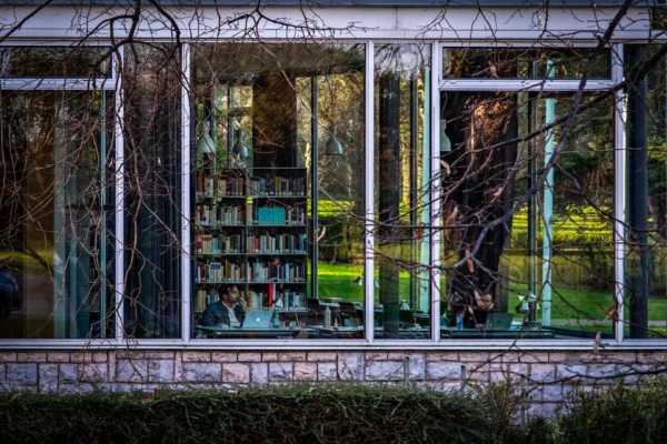 AnJo · 17/01/2026 · 1920 x 1280 Cette photographie montre une bibliothèque vue à travers une grande fenêtre vitrée. Le sujet principal est l’étagère remplie de livres colorés, principalement verts et bleus.La composition est symétrique, avec un cadrage serré sur la fenêtre encadrée par des montants rouges. La lumière naturelle, tamisée, crée des reflets et des contrastes doux. Les couleurs dominantes sont les teintes vertes et bleues des livres, avec des touches de gris et de brun en arrière-plan.Le style est épuré et réaliste, évoquant une ambiance calme et studieuse, presque intimiste. Les arbres dénudés ajoutent une touche mélancolique et hivernale.