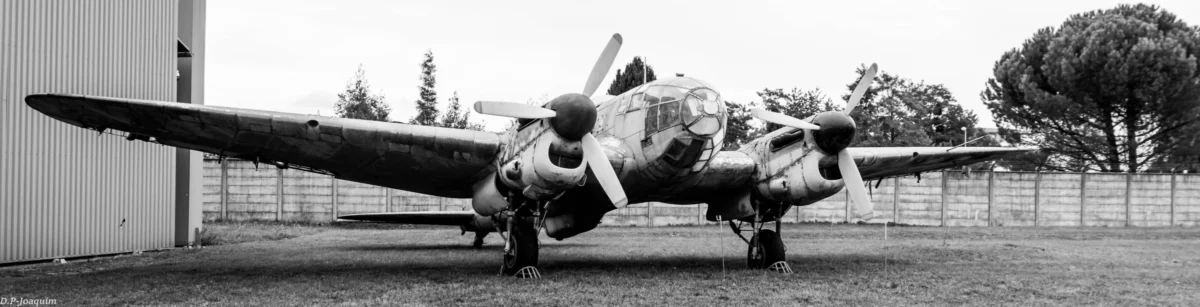 Pierre-Joaquim · 14/04/2015 · 6000 x 1532 Cette photographie en noir et blanc met en scène un avion de chasse ancien, sujet principal, posé sur une aire herbeuse. Le cadrage serré montre l’appareil sous différents angles, avec un pilote assis sur l’aile, ajoutant une touche humaine.La composition est dynamique, avec un jeu d’ombres et de lumière naturelle, soulignant les détails techniques de l’avion. Les tons gris et noirs dominent, accentuant le côté rétro et industriel. Le style documentaire, sobre et réaliste, crée une ambiance nostalgique, évoquant l’histoire de l’aviation militaire.