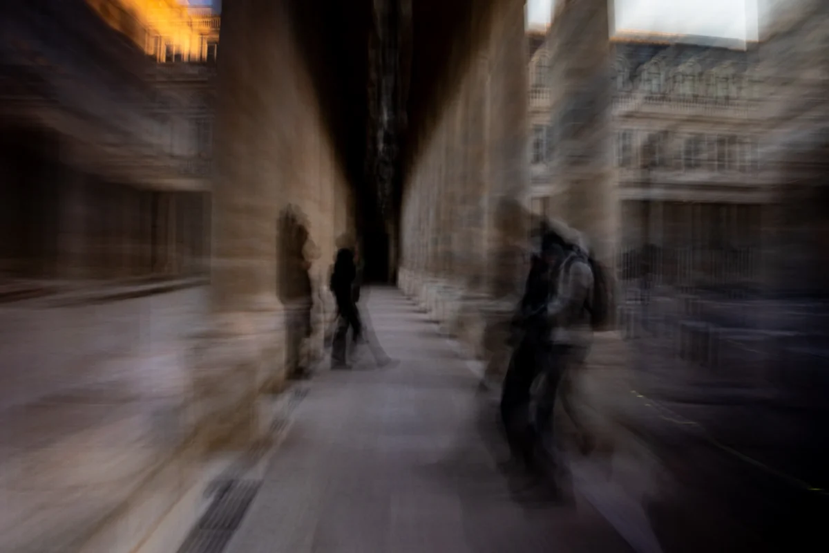 Jean-Marc · 03/01/2026 · 5712 x 3808 Cette photographie capture une scène floue et dynamique dans une ruelle urbaine. Le **sujet principal** semble être les silhouettes en mouvement, suggérant une vitesse ou un flou artistique. La **composition** est centrée sur une allée étroite bordée de bâtiments anciens, créant un effet de profondeur.Le **cadrage** est serré, avec une perspective légèrement inclinée. La **lumière** est tamisée, avec des teintes chaudes en arrière-plan, évoquant une ambiance nocturne ou crépusculaire. Les **couleurs dominantes** sont des tons grisâtres et dorés, renforçant l'atmosphère mystérieuse et cinétique. Le **style** est expressif et moderne, proche du flou d'action. L'**ambiance** est à la fois dynamique et mélancolique.