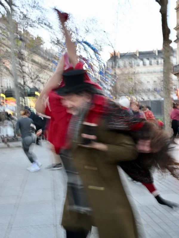 Xavier · 30/10/2025 · 3072 x 4096 Cette photographie capture une scène dynamique en mouvement flou. Le sujet principal semble être une personne en train de danser ou de célébrer, vêtue d’un haut rouge et d’un pantalon clair, avec un accessoire coloré à la main.La composition est déséquilibrée, avec un cadrage serré sur le sujet en mouvement. La lumière est naturelle, probablement en journée, mais le flou donne une impression de vitesse et d’énergie. Les couleurs dominantes sont les tons froids (gris du sol) et chauds (rouge du vêtement, bleu et blanc de l’accessoire).Le style est spontané et documentaire, évoquant une ambiance festive et joyeuse.
