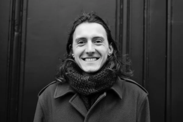 Daniel · 10/01/2026 · 3600 x 2400 Cette photographie met en scène un jeune homme souriant comme sujet principal. Le cadrage est serré, centré sur son visage et sa partie supérieure du torse. La composition est simple et symétrique, avec un fond uni et sombre.La lumière est douce et naturelle, éclairant le visage sans créer d’ombres marquées. Les couleurs dominantes sont en noir et blanc, ce qui accentue les contrastes et les textures, comme le pull et le foulard.Le style est épuré et intemporel, évoquant une ambiance à la fois moderne et classique, avec une touche chaleureuse grâce au sourire du sujet.