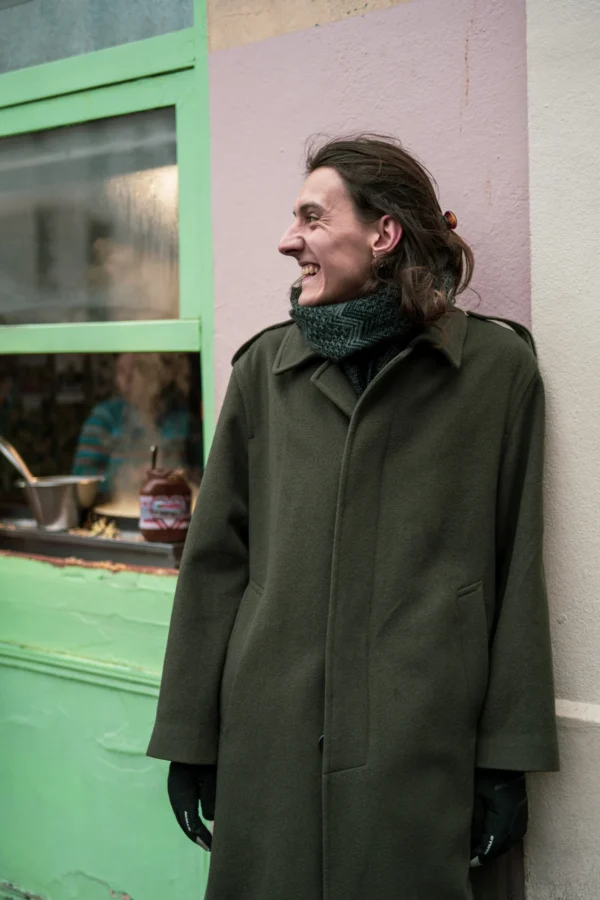 Daniel · 10/01/2026 · 2400 x 3600 Cette photographie montre un homme au regard pensif, sujet principal, vêtu d’un long manteau vert foncé et d’un foulard. Le cadrage est serré, centré sur lui, avec une porte verte en arrière-plan reflétant des éléments urbains.La lumière est naturelle et douce, créant des contrastes subtils. Les couleurs dominantes sont les verts (manteau, porte) et les tons neutres (visage, vêtements). Le style est sobre et réaliste, évoquant une ambiance calme et introspective, presque mélancolique.
