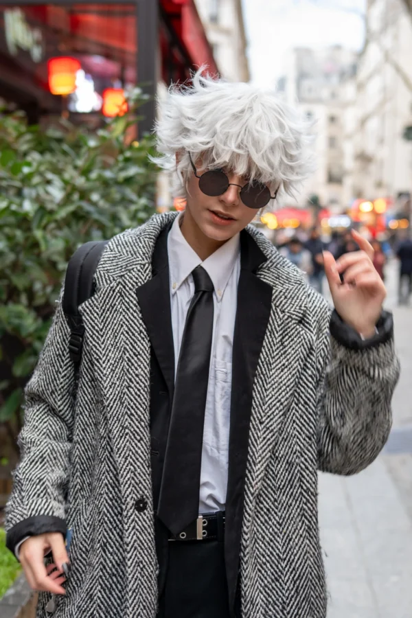 Daniel · 10/01/2026 · 2400 x 3600 Cette photographie met en scène un homme au look excentrique, sujet principal. Il arbore une perruque blanche hirsute, des lunettes noires, un long manteau à motifs et un nœud papillon, créant un style androgyne et décalé.La composition est dynamique, avec un cadrage serré sur le sujet, légèrement décentré. L’arrière-plan flou montre une rue animée, avec des éléments urbains et des passants.La lumière semble naturelle, douce, et les couleurs dominantes sont les tons gris du manteau, noir des lunettes et blanc de la perruque, renforçant un style rétro et avant-gardiste. L’ambiance est à la fois urbaine et artistique.