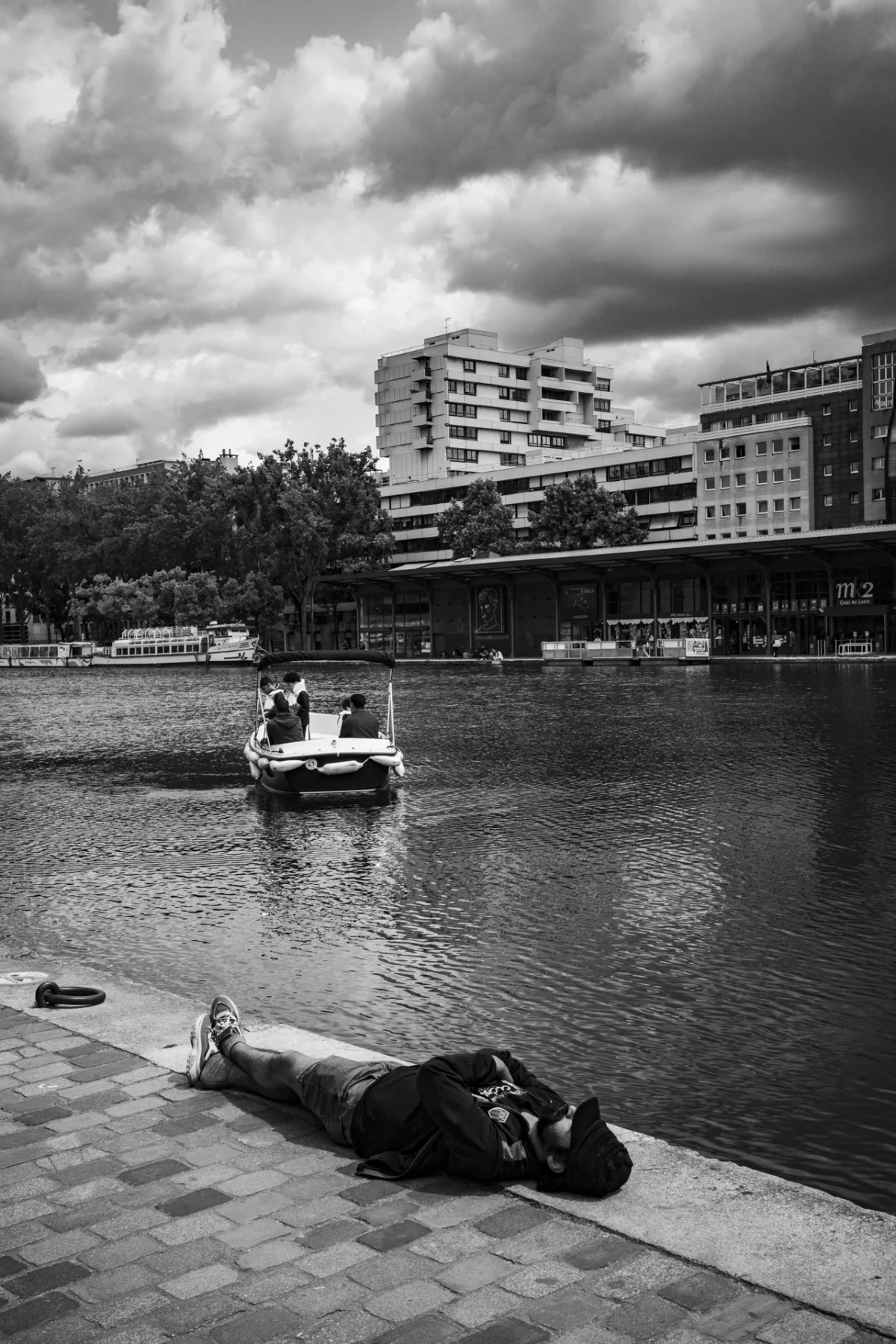 Pascal SC · 08/06/2025 · 1280 x 1920 Cette photographie en noir et blanc met en scène un homme allongé sur un trottoir en bordure d’eau, sujet principal. La composition oppose son immobilité à la dynamique du bateau en mouvement sur le lac. Le cadrage serré sur lui crée une intimité, tandis que l’arrière-plan urbain et les bâtiments ajoutent une dimension urbaine.La lumière, douce mais contrastée, accentue les ombres et les textures. Les tons dominants, du noir au gris clair, renforcent une ambiance mélancolique et contemplative. Le style, sobre et documentaire, évoque une scène de solitude en milieu urbain.