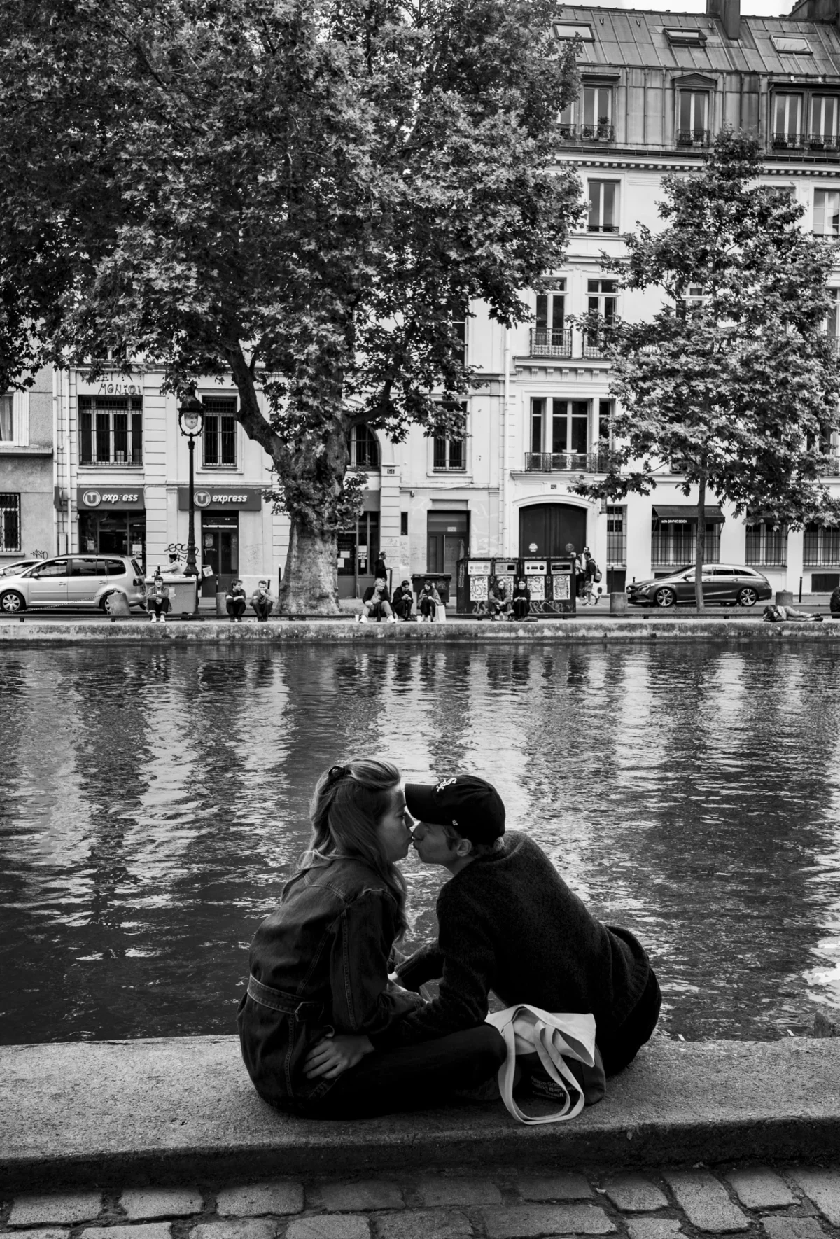 Pascal SC · 08/06/2025 · 1302 x 1920 Cette photographie en noir et blanc met en scène deux personnes assises sur une berge, partageant un moment intime. Le sujet principal est ce couple, cadré en premier plan, avec une composition qui les isole du reste.L’arrière-plan montre un bassin et des bâtiments classiques, créant une ambiance urbaine paisible. La lumière douce, probablement naturelle, adoucit les contours et accentue les contrastes. Le style est documentaire et romantique, évoquant une atmosphère mélancolique et introspective. Les tons dominants, en niveaux de gris, renforcent l’émotion et la nostalgie.