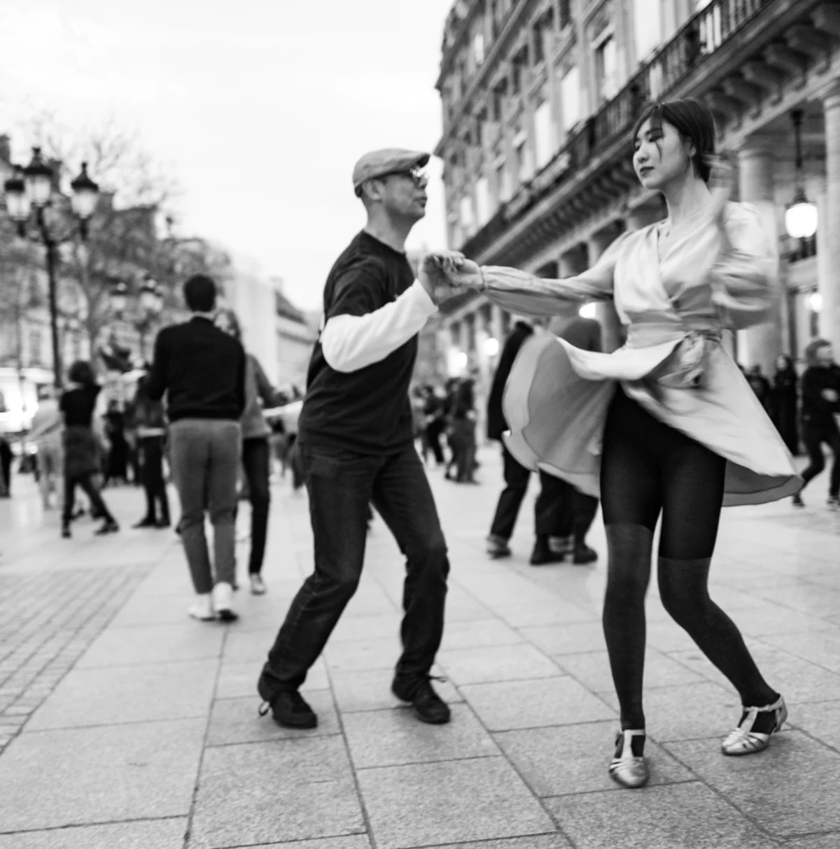 Pascal SC · 23/02/2025 · 1898 x 1920 Cette photographie en noir et blanc met en scène un couple comme sujet principal, dansant sur une rue animée. La composition est dynamique, centrée sur leurs mouvements synchronisés. Le cadrage serré les isole partiellement des passants en arrière-plan.La lumière est naturelle, douce, avec des contrastes modérés. Les couleurs dominantes, bien que monochromes, jouent sur les nuances de gris, renforçant l'atmosphère.Le style est documentaire et spontané, capturant un moment authentique. L’ambiance est joyeuse et vivante, évoquant une scène urbaine pleine d'énergie et de légèreté.