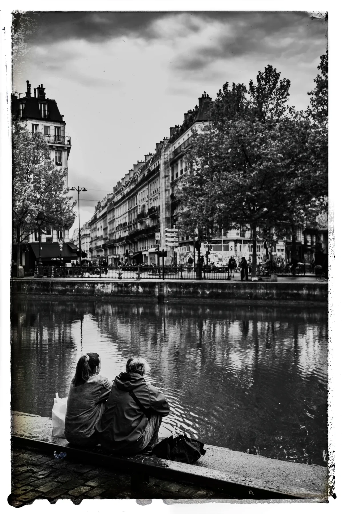 Pascal SC · 21/04/2025 · 1280 x 1920 Cette photographie en noir et blanc met en scène deux personnes assises sur une berge, dos à nous, observant un canal. Le sujet principal est leur posture contemplative, reflétée dans l’eau calme.La composition est équilibrée, avec le canal au premier plan et des bâtiments alignés en arrière-plan. Le cadrage est serré sur les personnages, encadrés par les arbres et les façades.La lumière douce et diffuse crée une ambiance paisible et mélancolique. Les tons dominants, en niveaux de gris, renforcent l’aspect intemporel et nostalgique. Le style rappelle le photojournalisme classique ou documentaire.