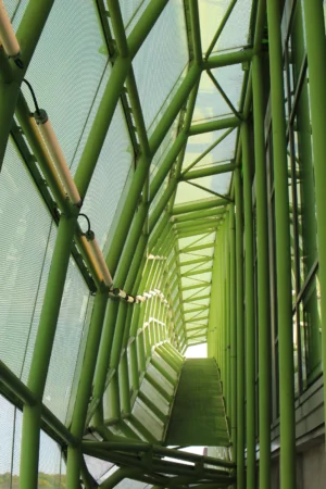 Cette photographie montre une structure architecturale moderne composée de tubes verts et de verre. Le sujet principal est l'escalier métallique central, encadré par des colonnes verticales en forme de bambou. La composition est géométrique et symétrique, avec un cadrage serré qui accentue les lignes et les perspectives.La lumière naturelle traverse les vitres, créant des jeux d'ombres et de transparence. Les couleurs dominantes sont le vert métallique des colonnes et le bleu-vert du verre, évoquant une ambiance futuriste et épurée. Le style est minimaliste et industriel, conférant une impression de modernité et de légèreté.