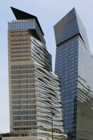 Cette photographie met en scène deux gratte-ciels modernes aux architectures distinctes. Le sujet principal est ces bâtiments, l’un avec une façade en damier et une pointe angulaire, l’autre en verre bleuté et courbes élancées.Le cadrage est serré, mettant en valeur leur hauteur et leurs détails architecturaux. La lumière, naturelle et douce, éclaire uniformément les structures, accentuant leurs lignes et textures. Les couleurs dominantes sont les tons gris, beige et bleu métallisé, reflétant un style contemporain et urbain.L’ambiance est à la fois dynamique et sophistiquée, évoquant une ville moderne et futuriste.