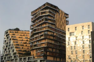 Cette photographie met en scène trois bâtiments modernes aux façades en bois et en béton. Le sujet principal est l’architecture contemporaine avec ses balcons et ses jeux de volumes.La composition est symétrique et verticale, avec un cadrage serré qui accentue la hauteur des édifices. La lumière, naturelle et douce, semble provenir d’un ciel légèrement voilé, créant des ombres subtiles.Les couleurs dominantes sont des tons bois chauds et des gris-beige, renforçant un style épuré et minimaliste. L’ambiance est à la fois urbaine et apaisante, reflétant une modernité architecturale harmonieuse.