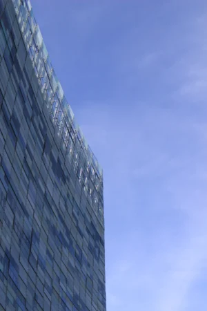 Cette photographie met en scène un **gratte-ciel moderne** recouvert de verre, sujet principal. La **composition** est épurée, avec un **cadrage serré** sur une partie de la façade, accentuant les reflets et les lignes verticales. La lumière, naturelle et douce, crée des jeux de transparence et de réflexion sur les vitres. Les **couleurs dominantes** sont le bleu du ciel et les teintes bleutées et grises du bâtiment.Le style est **minimaliste et contemporain**, évoquant une ambiance **urbaine et futuriste**, presque abstraite par son angle et ses reflets.