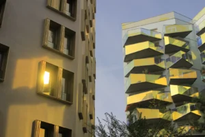 Cette photographie montre deux immeubles modernes en ville. Le sujet principal est l’architecture contrastée des bâtiments : l’un sobre et clair, l’autre aux balcons colorés en jaune et aux formes géométriques marquées.La composition est verticale, avec un cadrage serré mettant en valeur les façades. La lumière, douce et légèrement bleutée, suggère un coucher de soleil ou un crépuscule. Les couleurs dominantes sont les tons neutres (beige, gris) et les jaunes vifs des balcons.Le style est urbain et contemporain, évoquant une ambiance moderne et dynamique.