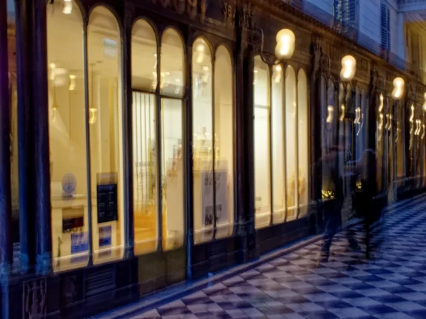 Cyrille · 03/01/2026 · 1200 x 900 Cette photographie montre une façade de boutique éclairée la nuit. Le sujet principal est la vitrine aux grandes baies vitrées, reflétant une lumière chaude et jaune.La composition est symétrique, avec des arcs architecturaux et des lampadaires alignés. Le cadrage est serré, mettant en valeur les détails des vitrines et du sol pavé en damier.Les couleurs dominantes sont les tons chauds (jaunes, ors) contrastant avec les teintes grises et noires de la nuit. Le style est urbain et élégant, créant une ambiance chaleureuse et intimiste malgré l'absence de personnes.