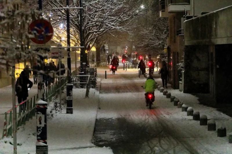 Cyrille · 05/01/2026 · 1500 x 997 Cette photographie capture une scène hivernale urbaine nocturne. Le **sujet principal** est un cycliste en tenue fluo, se déplaçant sur une piste enneigée.La **composition** montre une allée bordée de rails et de bâtiments, avec plusieurs personnes en mouvement. Le **cadrage** est serré sur le cycliste, mais inclut aussi l’arrière-plan pour donner une impression de profondeur.La **lumière** est tamisée, avec des éclairages artificiels et une ambiance nocturne. Les **couleurs dominantes** sont les tons froids (blanc de la neige, gris des bâtiments) adoucis par des éclats de rouge et de jaune (vêtements, lumières).Le **style** est réaliste et documentaire, créant une **ambiance** calme et légèrement mélancolique.
