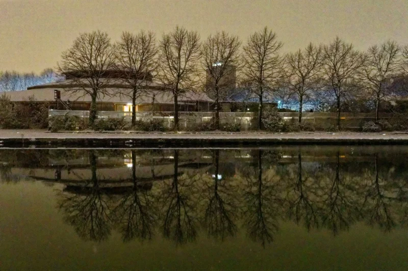 Cyrille · 05/01/2026 · 1200 x 798 Cette photographie met en scène un bâtiment traditionnel reflété dans une étendue d’eau calme. Le sujet principal est ce pavillon et son reflet, créant une symétrie harmonieuse. La composition est équilibrée, avec une ligne d’eau centrale.Le cadrage est large, capturant aussi des arbres et un ciel légèrement teinté. La lumière, douce et tamisée, suggère un coucher de soleil ou un éclairage artificiel chaud. Les couleurs dominantes sont des tons verts, bruns et jaunes dorés, renforçant une ambiance paisible et sereine. Le style est épuré et contemplatif, évoquant une scène presque onirique et intemporelle.