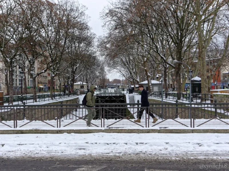 Cyrille · 07/01/2026 · 1200 x 900 Cette photographie capture une scène hivernale urbaine. Le **sujet principal** est un groupe de personnes traversant un pont recouvert de neige légère. La **composition** est symétrique, avec le pont central et les arbres dénudés de chaque côté.Le **cadrage** est large, incluant une partie de la route et des bâtiments en arrière-plan. La **lumière** est douce et diffuse, typique d’un jour d’hiver nuageux. Les **couleurs dominantes** sont les tons froids : blanc de la neige, gris des arbres et du ciel, avec des touches de brun et de noir pour les structures.Le **style** est réaliste et documentaire, évoquant une **ambiance** calme et paisible, presque mélancolique.