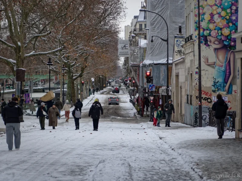 Cyrille · 07/01/2026 · 1200 x 900 Cette photographie capture une rue enneigée en ville. Le **sujet principal** est la vie urbaine sous la neige, avec des **piétons** marchant sur un trottoir glacé.La **composition** est linéaire, avec une perspective en profondeur accentuée par les bâtiments et les arbres bordant la rue. Le **cadrage** est serré, mettant en valeur les détails des passants et de l’architecture.La **lumière** est naturelle mais tamisée, typique d’un jour d’hiver. Les **couleurs dominantes** sont les tons froids : blanc de la neige, gris des bâtiments, et quelques touches de couleurs vives sur les murs et vêtements.Le **style** est documentaire et réaliste, évoquant une **ambiance** paisible et quotidienne, malgré le froid.