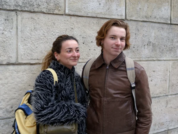 Cyrille · 10/01/2026 · 2000 x 1500 Cette photographie montre deux jeunes posant côte à côte contre un mur en pierre.Le sujet principal est les deux personnes, un homme et une femme souriants. La composition est symétrique, avec un cadrage serré sur eux. La lumière semble naturelle, douce et légèrement tamisée, créant des ombres peu marquées. Les couleurs dominantes sont les tons neutres (beige, gris) du mur et des vêtements, avec des touches de bleu et de marron.Le style est simple et spontané, évoquant une ambiance détendue et amicale.