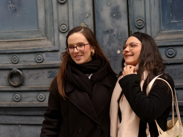 Cyrille · 10/01/2026 · 2000 x 1500 Cette photographie montre deux jeunes femmes souriantes posant devant une porte en métal ouvragé. Le sujet principal est elles, en premier plan, avec une composition serrée et naturelle. Le cadrage est horizontal et centré sur leurs visages.La lumière semble douce et naturelle, probablement en extérieur par temps couvert. Les couleurs dominantes sont les tons froids (gris de la porte, noir des vêtements) et quelques touches chaudes (teintes des écharpes et des visages).L’ambiance est chaleureuse et conviviale, avec un style simple et spontané.