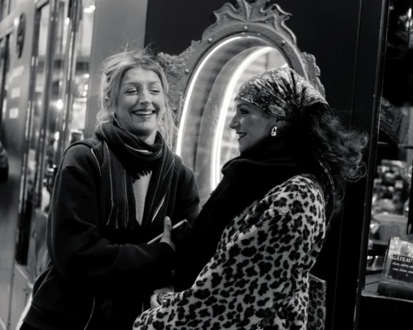 Cyrille · 10/01/2026 · 2000 x 1600 Cette photographie en noir et blanc met en scène deux femmes comme sujets principaux. La composition montre une interaction amicale devant un miroir orné. Le cadrage est serré, centré sur elles, avec un arrière-plan flou.La lumière semble naturelle, douce et légèrement tamisée. Les couleurs dominantes, bien que réduites au noir et blanc, suggèrent des tons sombres et contrastés.Le style évoque une élégance rétro, renforcée par les accessoires et les tenues sophistiquées. L’ambiance est chaleureuse et intime, mêlant humour et complicité.