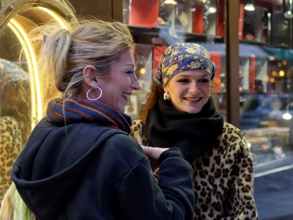 Cyrille · 10/01/2026 · 2000 x 1500 Cette photographie montre deux jeunes femmes comme sujets principaux, souriantes et en conversation. La composition est serrée, centrée sur elles, avec un cadrage en plan moyen. L’ambiance semble chaleureuse et détendue.La lumière est naturelle, probablement en journée, avec des ombres douces. Les couleurs dominantes incluent des tons neutres (beige, noir) et des touches vives (violet du bandeau, orange des accessoires). Le style est spontané et réaliste, évoquant une scène de rue animée.