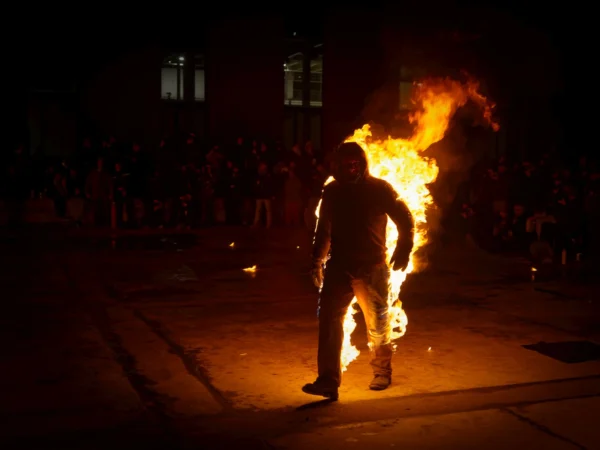 Cyrille · 24/01/2026 · 2026 x 1520 Cette photographie met en scène un homme comme sujet principal, manipulant une torche enflammée dans une rue sombre. La composition est dynamique, centrée sur l'individu et son feu, avec un cadrage serré qui isole la scène.Les lumières sont contrastées : l'éclat vif du feu domine, tandis que l'arrière-plan reste plongé dans l'obscurité. Les couleurs dominantes sont les teintes chaudes (oranges, jaunes) du feu, contrastant avec les tons sombres de la nuit.Le style est réaliste et brut, évoquant une ambiance intense et mystérieuse, presque théâtrale.