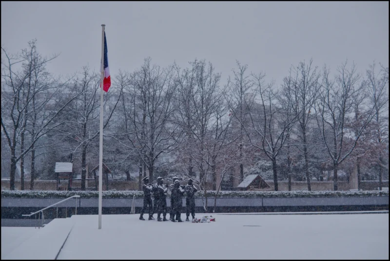 Frédéric · 05/01/2026 · 2510 x 1680 Cette photographie montre un groupe de personnes, probablement des militaires, alignées sous un drapeau français enneigé. Le sujet principal est le drapeau et les individus en uniforme.La composition est symétrique, avec le groupe centré sous le drapeau. Le cadrage est large, incluant un arrière-plan de arbres dénudés et un ciel gris.La lumière est diffuse, typique d’un jour d’hiver, créant une ambiance froide et sobre. Les couleurs dominantes sont les tons blancs de la neige, les gris du ciel et des arbres, et le bleu, blanc, rouge du drapeau. Le style est réaliste et documentaire, évoquant une ambiance solennelle et austère.