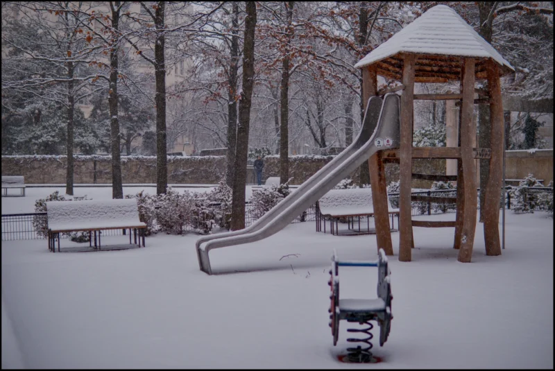Frédéric · 05/01/2026 · 2511 x 1680 Cette photographie montre un **parc d’attractions pour enfants** recouvert de neige. Le **sujet principal** est le toboggan et l’aire de jeu en bois partiellement ensevelis sous la neige.La **composition** est symétrique, avec un **cadrage large** incluant des arbres nus et un banc en arrière-plan. La **lumière** est douce et diffuse, typique d’un jour hivernal couvert. Les **couleurs dominantes** sont les tons froids : blanc de la neige, gris du ciel et brun des structures en bois.Le **style** est réaliste et documentaire, évoquant une **ambiance calme et mélancolique**, presque déserte, renforcée par le silence hivernal.