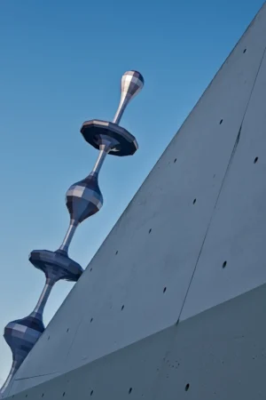Cette photographie montre un **sujet architectural moderne** : une série de **trois éléments en forme d’entonnoirs métalliques** fixés à une paroi inclinée et perforée. La **composition** est épurée, axée sur la verticalité et la géométrie. Le **cadrage serré** isole ces éléments contre un fond de ciel bleu clair, accentuant leur design minimaliste.La **lumière**, naturelle et vive, met en valeur les textures métalliques et les jeux d’ombres. Les **couleurs dominantes** sont le gris anthracite des structures et le bleu intense du ciel, créant un contraste saisissant. Le style est **contemporain et industriel**, évoquant une ambiance **urbaine et futuriste**.