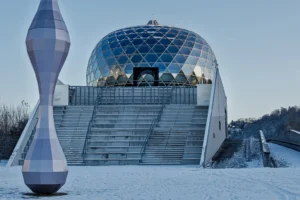 Cette photographie met en scène un bâtiment futuriste aux formes géométriques, dominé par une structure en dôme transparente et des lignes anguleuses. Le sujet principal est ce bâtiment, entouré de neige, ce qui accentue son aspect architectural innovant.La composition joue sur les contrastes entre les formes organiques et les angles droits. Le cadrage inclut une sculpture abstraite en premier plan, ajoutant une touche artistique. La lumière, douce et bleutée, crée une ambiance froide et mystérieuse. Les couleurs dominantes sont les tons gris, blancs et bleus, renforçant une atmosphère moderne et minimaliste. Le style est épuré et contemporain, évoquant une ambiance futuriste et presque onirique.