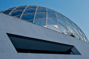 Cette photographie met en scène un bâtiment moderne avec une structure géométrique dominante : une coupole vitrée en forme de treillis métallique. Le cadrage est serré, centré sur cette partie architecturale, avec un angle légèrement bas.La lumière naturelle éclaire la scène, créant des contrastes nets entre les zones ombrées et lumineuses. Les couleurs dominantes sont le gris (béton, structure) et le bleu (ciel). Le style est épuré et contemporain, évoquant une ambiance futuriste et technologique. L’effet est à la fois minimaliste et impressionnant.